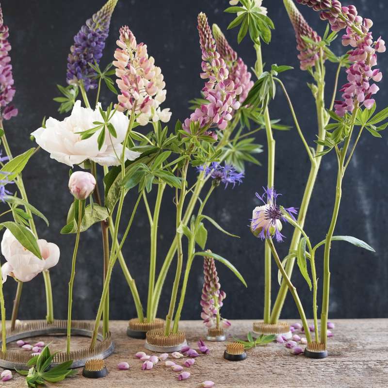 Assorted flowers, including lupines and roses, elegantly displayed on a 34mm Hanataba kenzan ring, set against a dark backdrop with scattered petals on the surface.