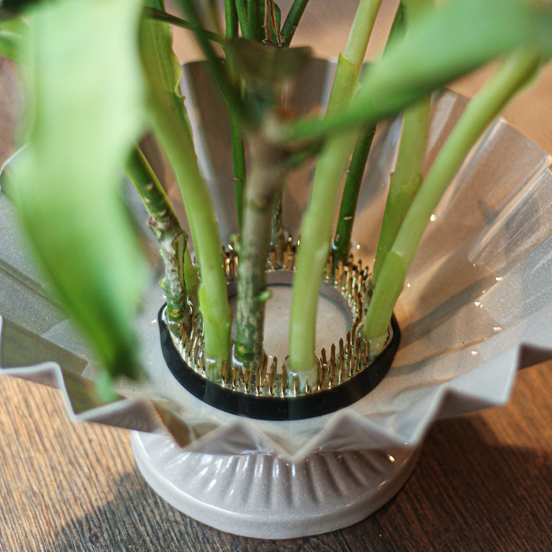 Nahaufnahme eines 70-mm-Kenzans an der Basis eines Blumenarrangements. Seine Messingstifte halten grüne Stängel sicher über einer geriffelten Glasschale und zeigen das wesentliche Werkzeug der Ikebana-Blumenkunst.