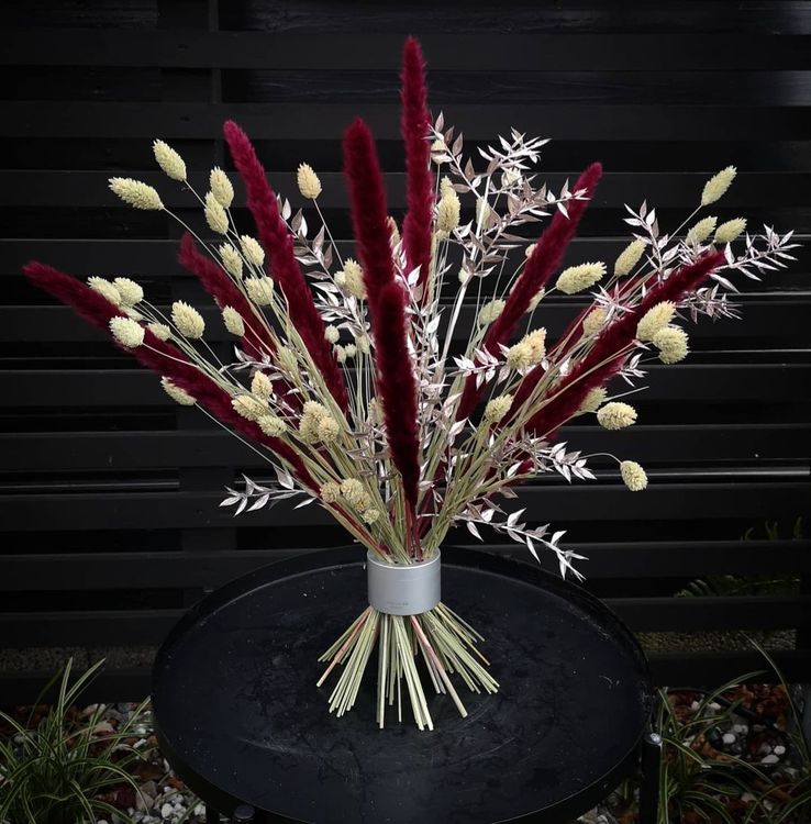 Ein lebhaftes Ikebana-Arrangement aus purpur- und cremefarbenen Trockenblumen in einem Hanataba-Bouquet-Twister vor einem Hintergrund aus schwarzen Latten