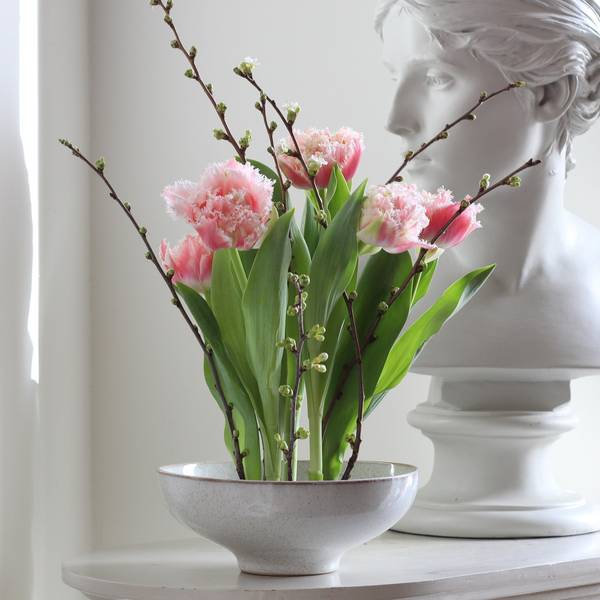 Elegantes Blumenarrangement mit rosa gefransten Tulpen und zarten knospenden Zweigen in einer gesprenkelten Schale, daneben eine klassische weiße Büste.