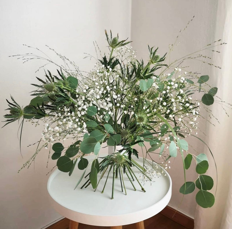 Elegantes Ikebana-Arrangement mit einer Auswahl an Grünzeug und weißen Blüten, das von einem Hanataba Bouquet Twister in Perlmuttsilber auf einem weißen Tisch gehalten wird.