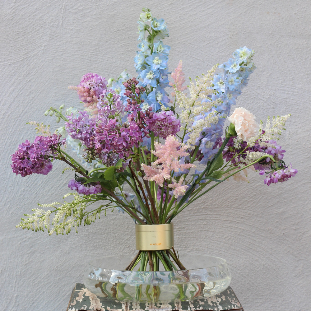 Elegantes Ikebana-Blumenarrangement mit einer Vielzahl von pastellfarbenen Blüten, darunter Flieder und Rittersporn, die von einem schicken Spiralstielhalter gehalten werden.