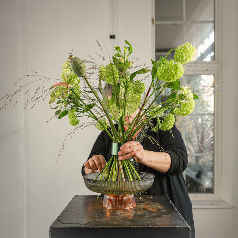 Ein Florist fertigt ein professionelles Blumenarrangement mit Viburnum-Schnittblumen in Hanataba Efeugrün an, das in einem gestreiften runden Glastablett präsentiert wird.