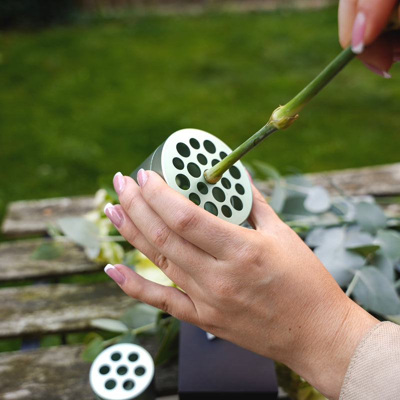 Eine Person mit rosafarbenem Nagellack arrangiert Eukalyptus und Blumen, indem sie einen Stiel in einen Hanataba Matcha Green Bouquet Twister über einem Holztisch im Garten steckt.