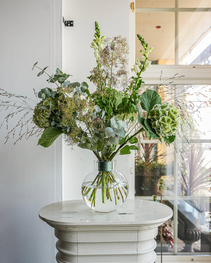 Eine klare Glasvase mit einem Arrangement aus grünen und weißen Blumen, darunter Hortensien und Eukalyptus, steht auf einem kleinen weißen runden Tisch. Der Strauß Hanataba Ivy Green sticht hervor, während das Sonnenlicht durch ein hohes Fenster im Hintergrund fällt.