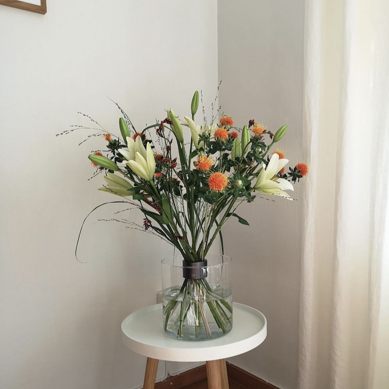 Minimalistisches Blumenarrangement im Ikebana-Stil mit weißen Lilien und orangefarbenem Carthamus in einer klaren Vase mit einem Hanataba-Spiralstielhalter auf einem weißen Tisch.