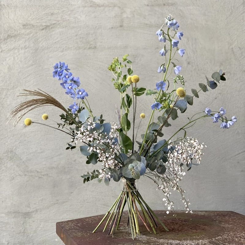 Ikebana-Blumengesteck mit blauen Rittersporn, gelben Craspedia und zartem Schleierkraut, arrangiert mit dem traditionellen Hanataba-Spiralstielhalter mit japanischen Techniken.