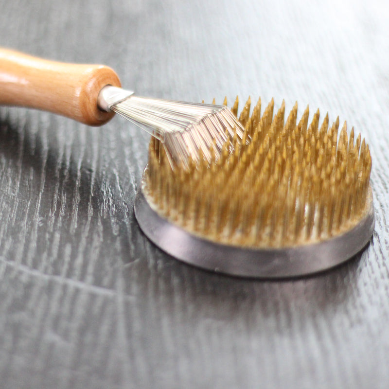 Eine Kenzan-Harke mit Holzgriff wird zum Durchkämmen der Messingnadeln eines goldenen Kenzan verwendet, der auf einer holzgetönten Oberfläche steht.