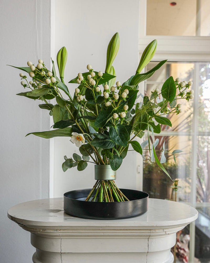 Wunderschönes Blumenarrangement mit Lilien auf einem schwarzen Blumentablett auf einem weißen Sockel in einem schönen weißen Raum mit getäfelten Fenstern.