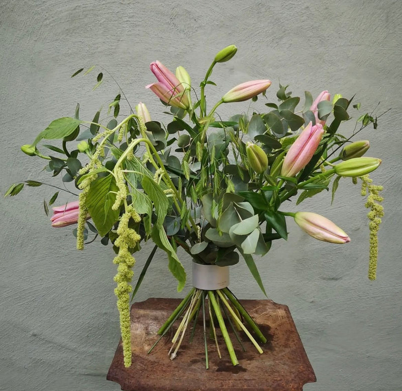 Ein üppiges Blumenarrangement mit rosafarbenen Lilien und kaskadenartigen grünen Akzenten, zusammengehalten von einem Hanataba Bouquet Twister in Perlmuttsilber, vor einer strukturierten grauen Wand