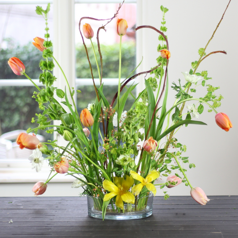 Ein lebhaftes Arrangement aus Tulpen und gemischtem Grün, das aufrecht in einem 200-mm-Kenzanring steht und auf einem dunklen Holztisch in der Nähe eines Fensters platziert ist.