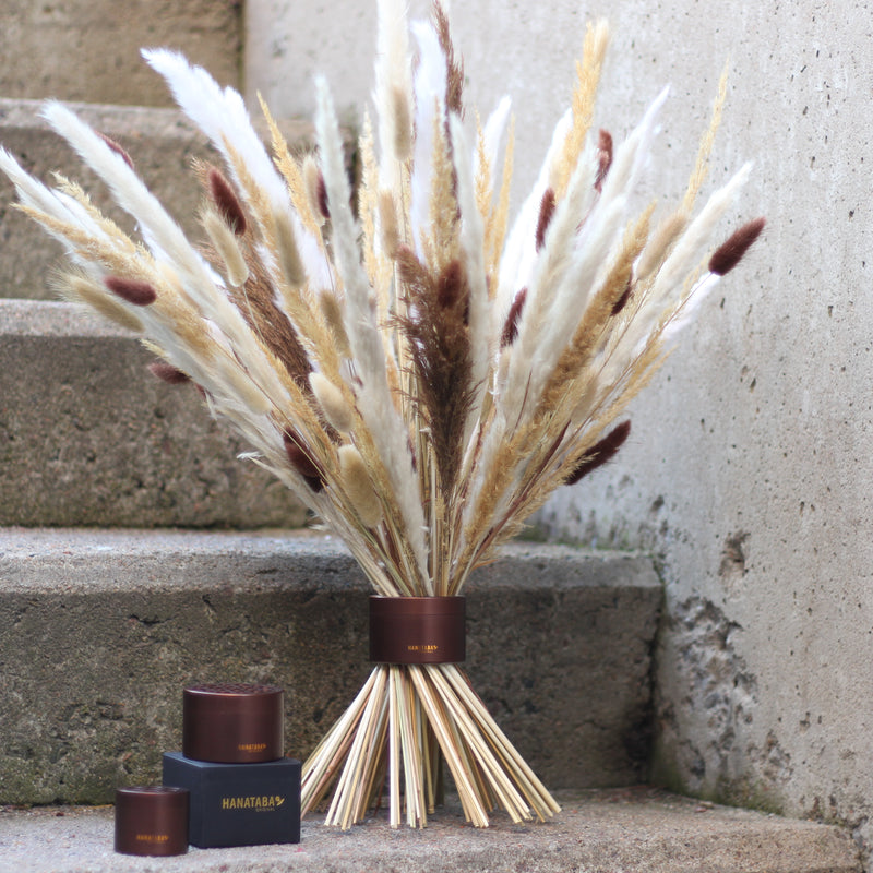 Ein Muddy Brown Hanataba Bouquet Twister stützt elegant ein Arrangement aus getrockneten Gräsern und Schilf auf Betonstufen, begleitet von zwei passenden Hanataba Behältern.