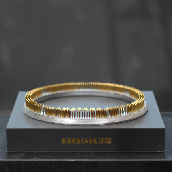 Ein glänzender 200-mm-Kenzan-Ring ruht auf einer eleganten grauen Präsentationsbox mit der goldenen Aufschrift "HANATABA 花束", die Eleganz und Funktionalität in einem Blumenarrangement präsentiert.