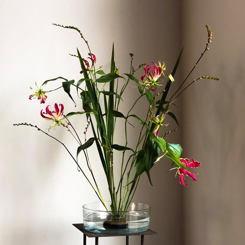 Exotische Gloriosa-Lilien mit auffallend rosafarbenen und gelben Blütenblättern, verankert durch einen 70-mm-Kenzan-Ring in einer Glasschale.