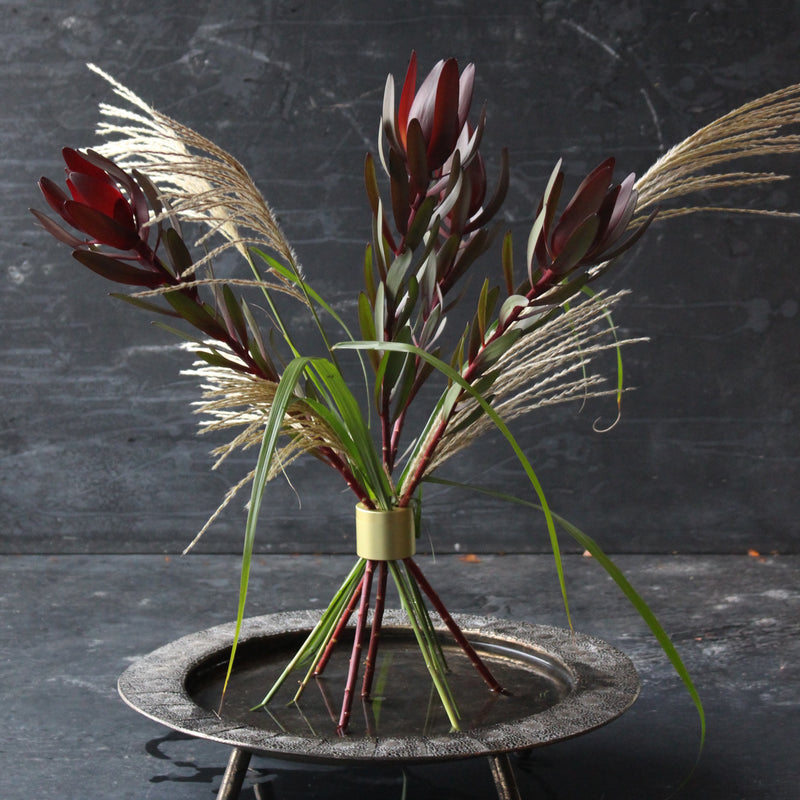 Ein geschmackvolles Blumenarrangement mit tief burgunderroten Leucadendronblüten und zarten Wildgräsern, elegant zusammengehalten von einer Hanataba in Champagnergold.