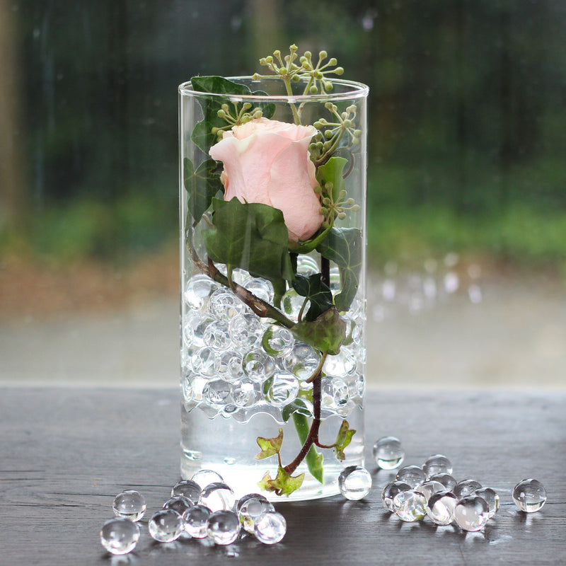 Eine einzelne blassrosa Rose mit Efeu in einer klaren Vase, gefüllt mit Wasser und transparenten Hydrogelperlen.