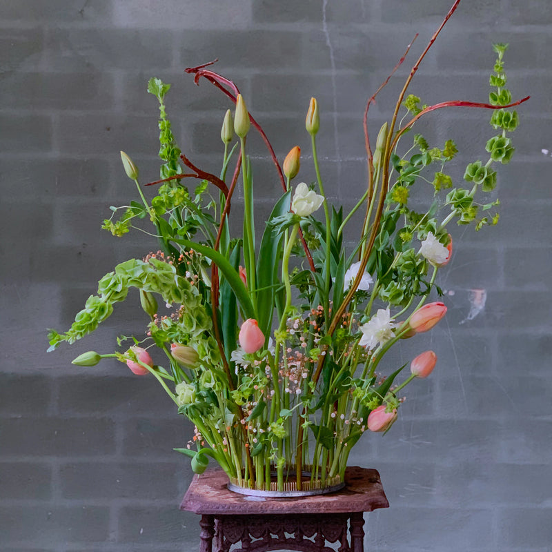 Ein lebhafter Strauß mit Tulpen und einer Reihe von Grünpflanzen, sorgfältig arrangiert mit einem 200-mm-Kenzan-Ring auf einem verzierten Holzsockel vor einer grauen Ziegelwand.