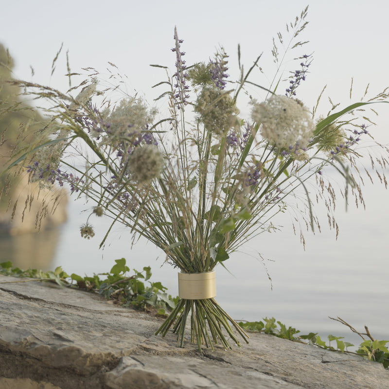 Wildblumen, darunter lilafarbene Ähren und weiße Büschel, wurden in einem champagnergoldenen Hanataba-Bouquet auf einem Felsvorsprung mit Blick auf das ruhige Wasser versammelt.