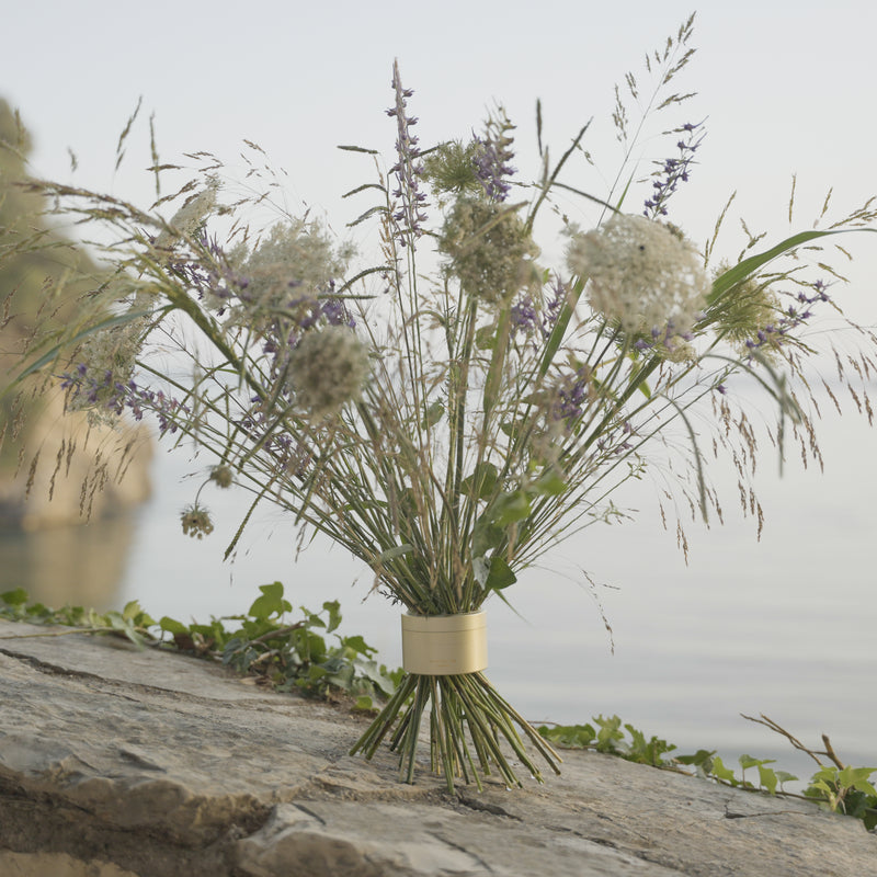 Wildblumen und Gräser in einem Hanataba Bouquet Twister Champagne Gold, das auf einem Felsen mit ruhigem Wasser im Hintergrund steht und natürliche Ruhe vermittelt.